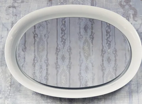 Ein ovaler Spiegel mit einem weißen Rahmen hängt vor einer strukturierten Tapete mit zarten, graublauen Mustern. 