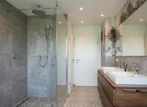 Großzügige Dusche mit grauen Feinsteinzeugfliesen und Glasabtrennung, daneben Doppelwaschtisch mit Spiegel.