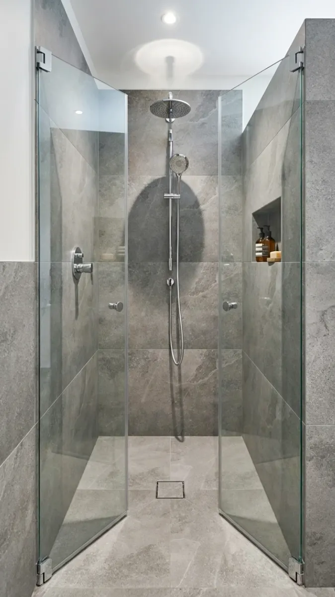 Begehbare Dusche mit Regendusche, grauen Fliesen und Nische für Pflegeprodukte.