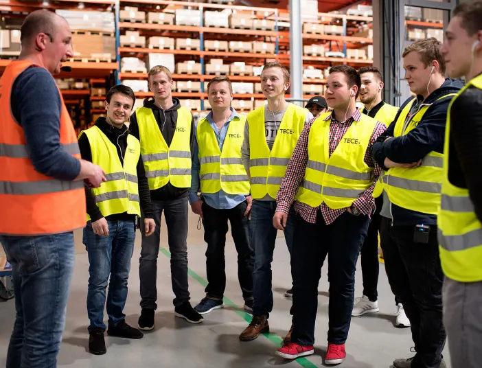 Mitarbeiter in gelben Sicherheitswesten nehmen an einer Führung durch ein großes Lager teil, geleitet von einem Mann in einer orangefarbenen Weste.